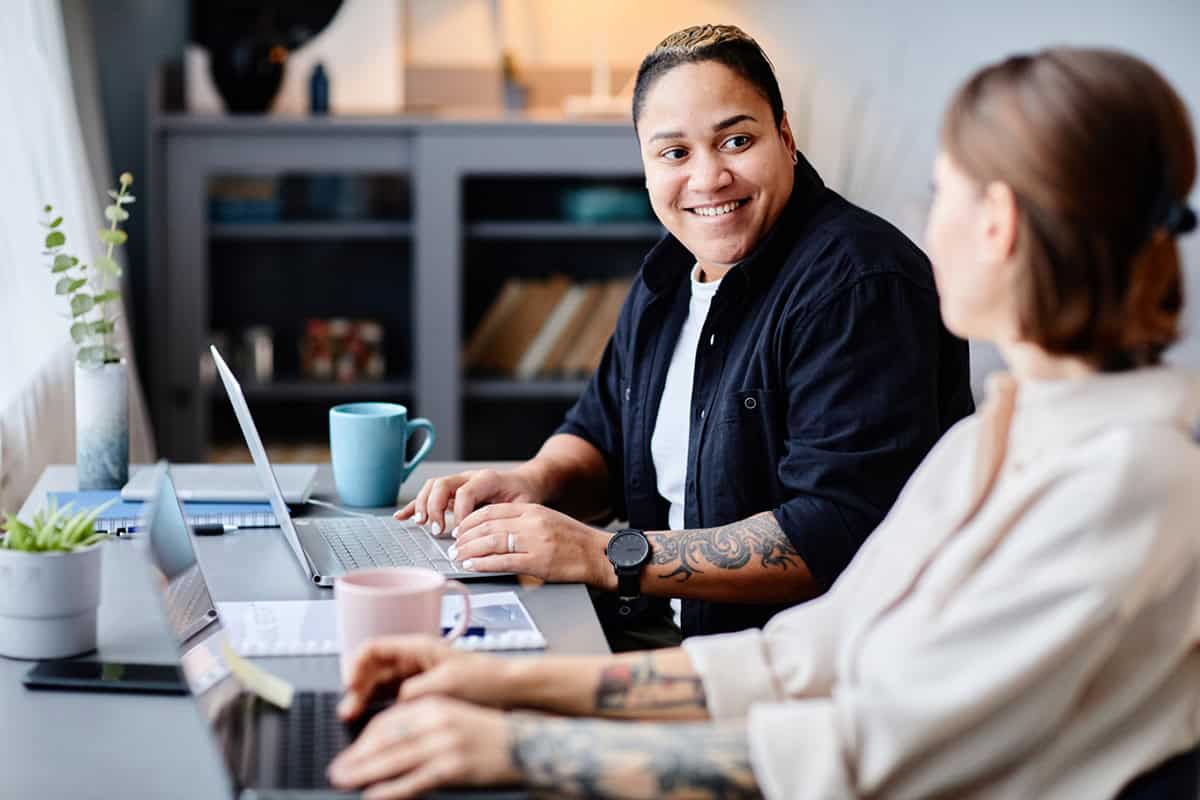 Two people sit at a desk with laptops, smiling and talking, with coffee mugs and papers nearby.