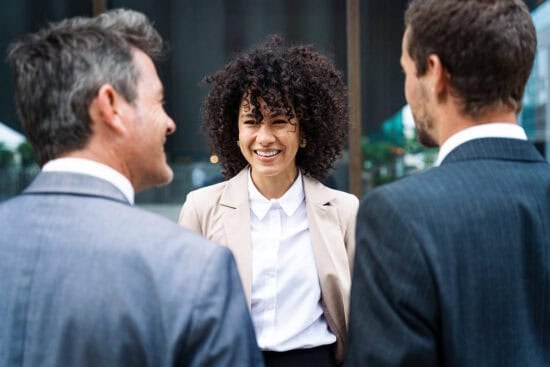 A smiling woman in business attire talks with two men in suits outdoors.