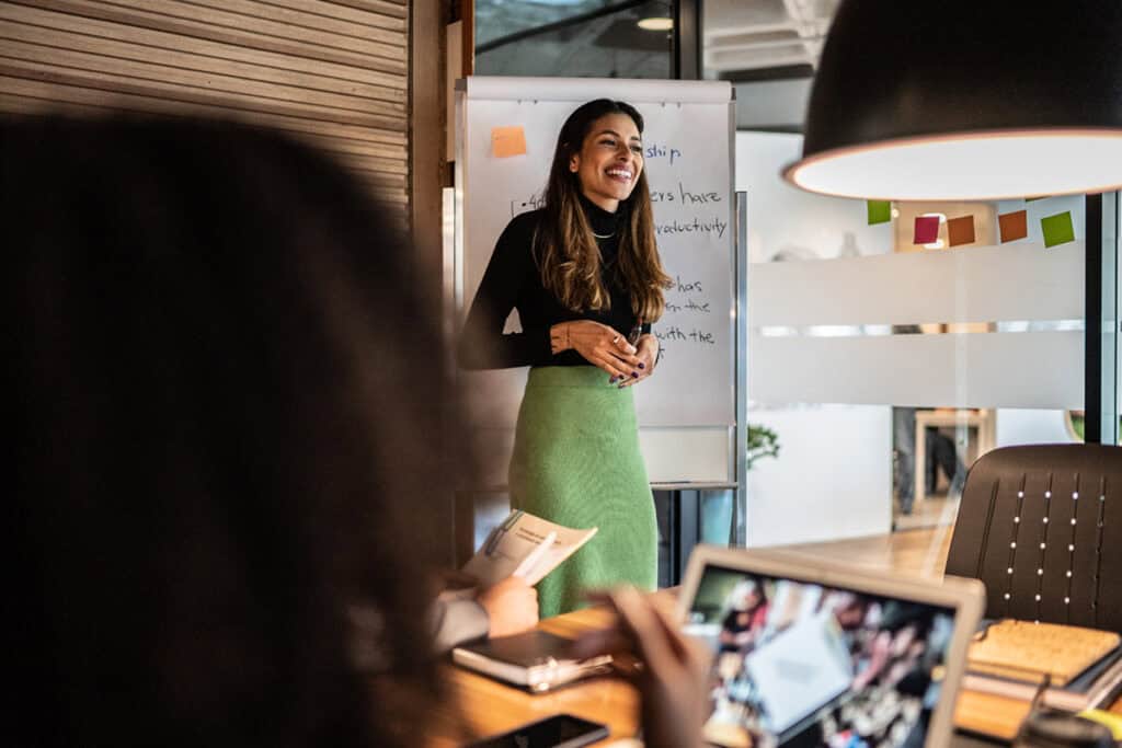 Woman smiling and presenting by a whiteboard in a modern office meeting room.