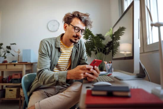 Man with glasses sitting at a desk, looking at his phone, with a computer and notebooks nearby.