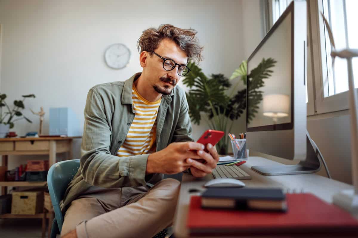 Man with glasses sitting at a desk, looking at his phone, with a computer and notebooks nearby.