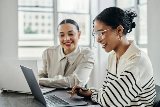 Two women smiling and working together on laptops at a desk in a bright, modern office.