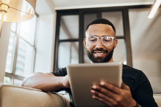 A man with glasses smiles while using a tablet, sitting on a couch in a bright, modern room.