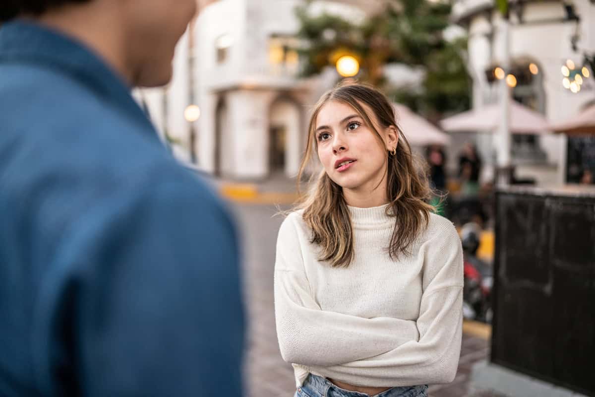 Young woman with crossed arms listens to person in casual outdoor setting, looking thoughtful.
