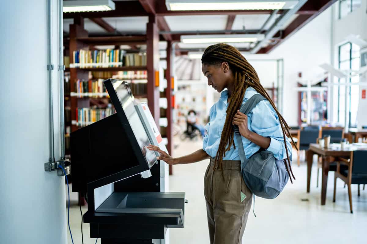 A woman uses a touchscreen kiosk in a library, wearing a backpack and standing near bookshelves.