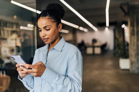 Woman in a blue shirt using a smartphone while standing in a modern office setting.