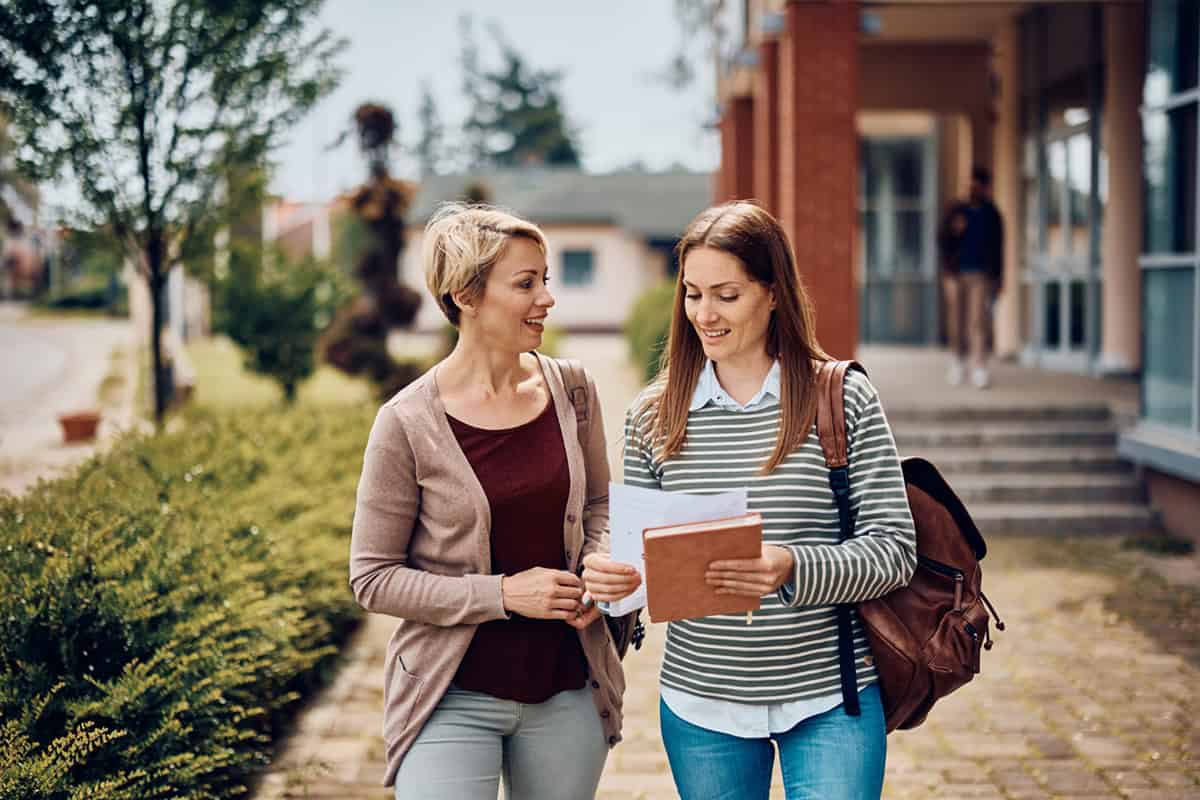 Two women walking and talking outside, one holding notebooks and wearing a backpack, in a campus setting.