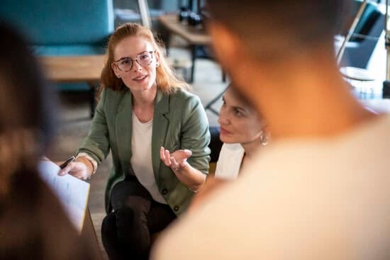 A woman in glasses speaks while holding papers in a casual meeting with colleagues.