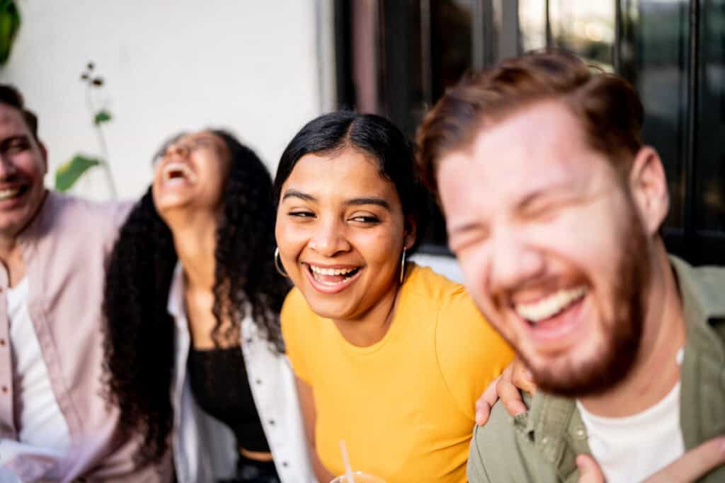 Four friends sitting close together, laughing and smiling outdoors on a sunny day.