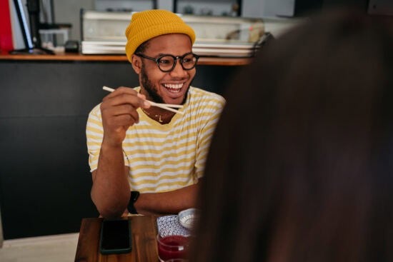 Smiling man in a yellow beanie holding chopsticks, sitting at a table with food in front of him.