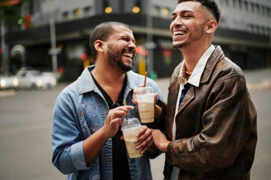 Two friends laughing together while holding iced coffee drinks on a city street.