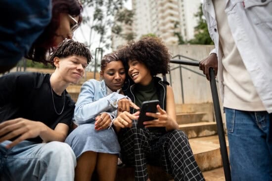 A group of friends sitting on outdoor steps, smiling and looking at a phone together.