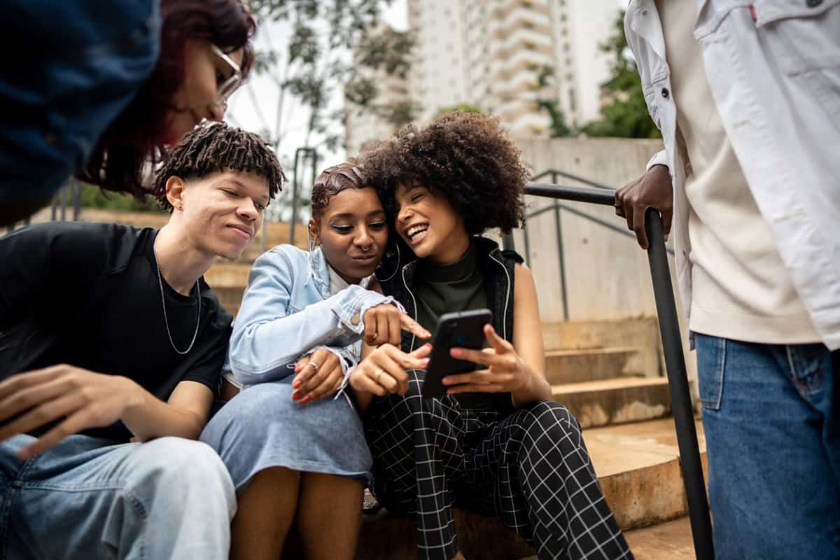 A group of friends sitting on outdoor steps, smiling and looking at a phone together.