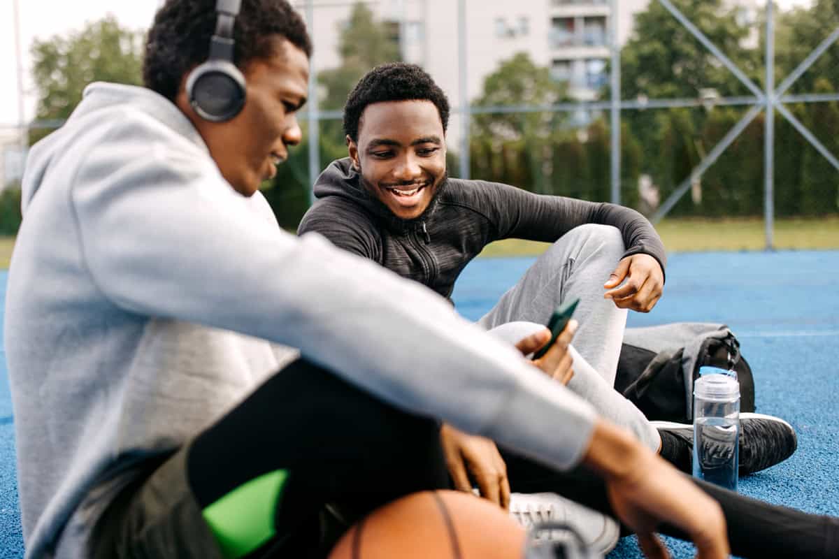 Two young men sitting on an outdoor basketball court, smiling and looking at a phone together.