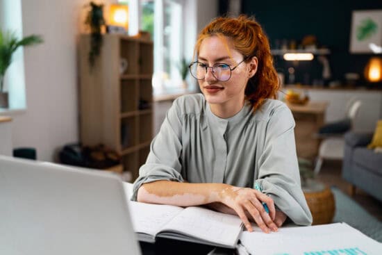 A woman with glasses studies at a desk with a laptop and open notebook in a bright, modern room.