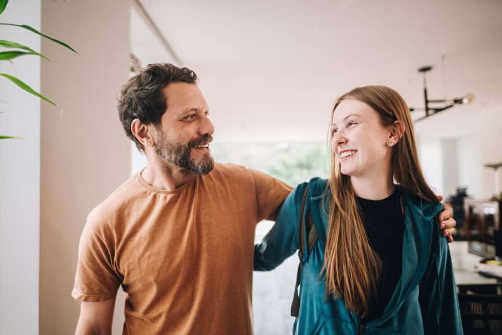 A man and a young woman smiling at each other indoors, with the man's arm around her shoulders.