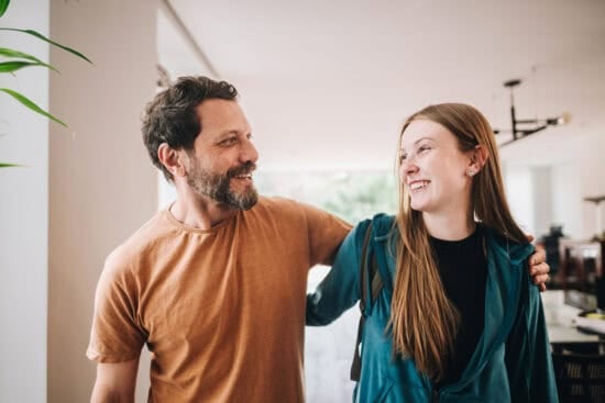 A man and a young woman smiling at each other indoors, with the man's arm around her shoulders.