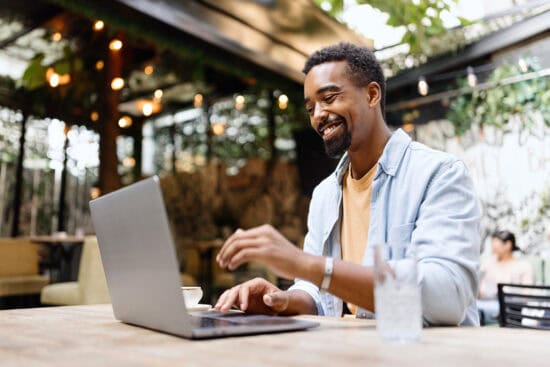 Smiling man uses a laptop at an outdoor cafe, with a glass of water and greenery in the background.