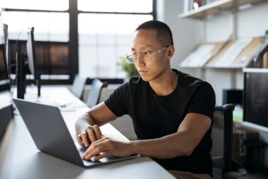 Man with glasses working on a laptop at a desk in a modern office with shelves and large windows.