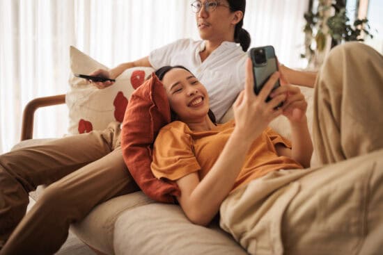 Two people relax on a couch; one smiles at their phone while the other holds a TV remote.