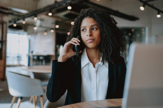 Woman in business attire sits at a table, talking on a phone, with a laptop in front of her.