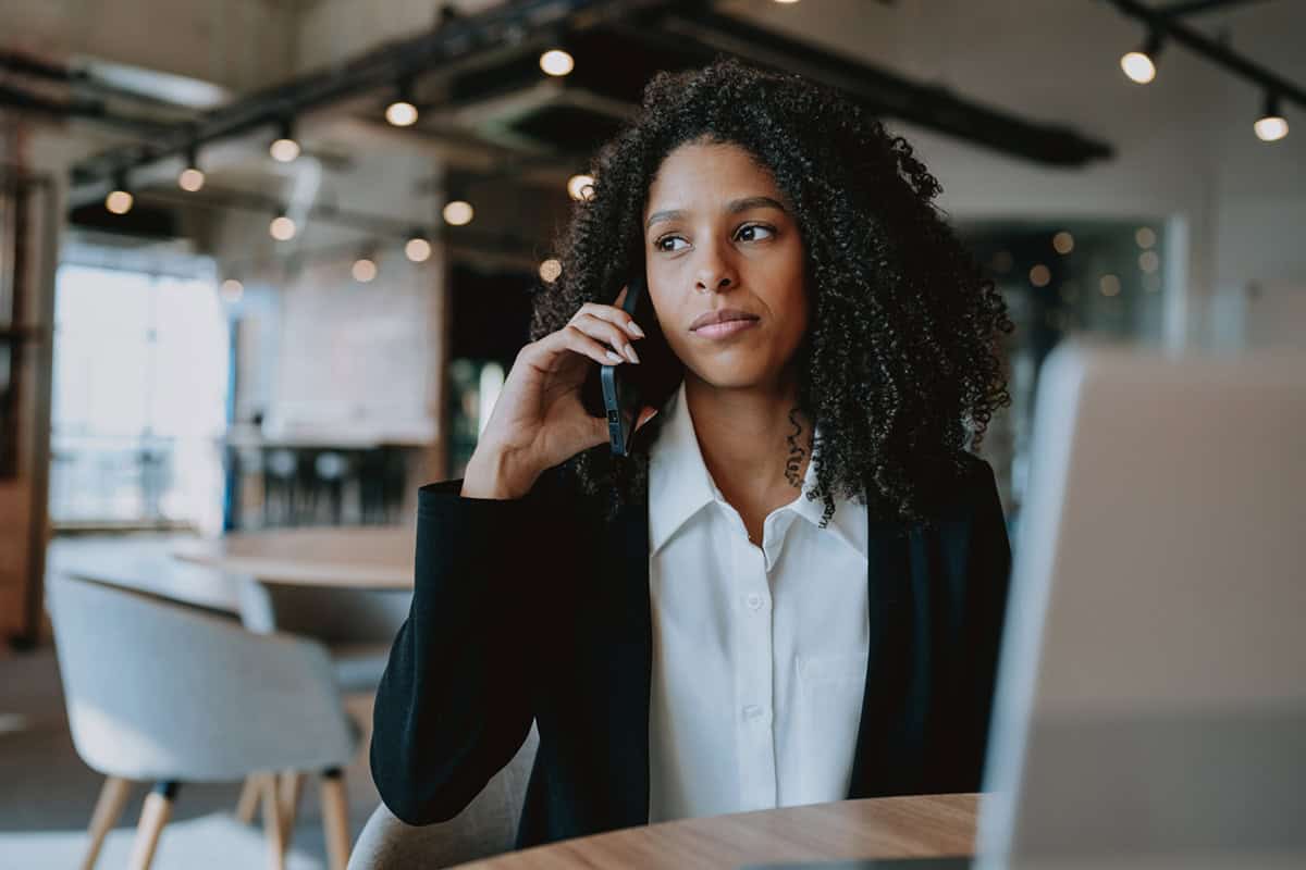 Woman in business attire sits at a table, talking on a phone, with a laptop in front of her.
