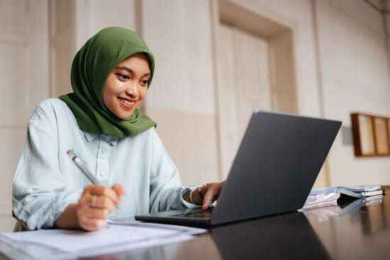 Smiling woman in a green hijab studies at a table with a laptop, notebook, and pen.