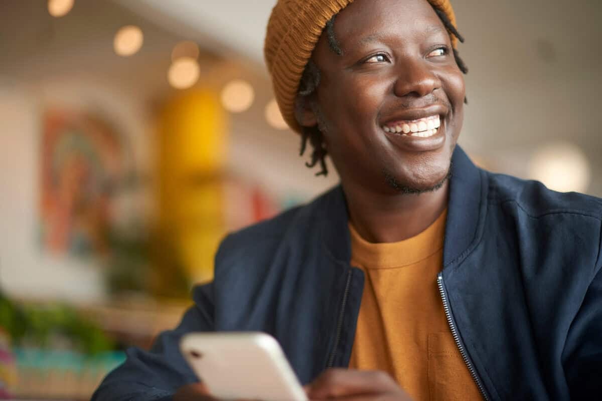 Smiling man in a brown beanie holding a smartphone, sitting indoors with a blurred colorful background.