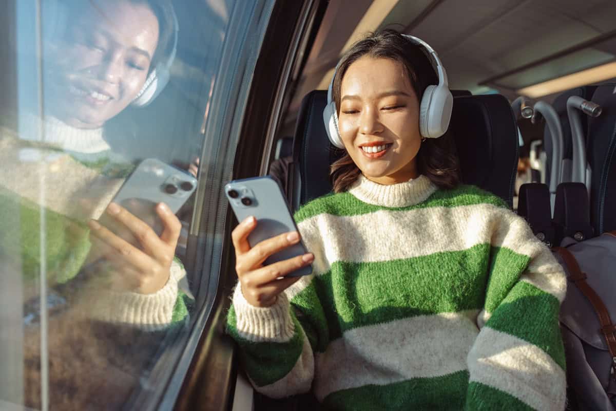 Woman wearing headphones smiles while looking at her smartphone on a train, sunlight shining through the window.