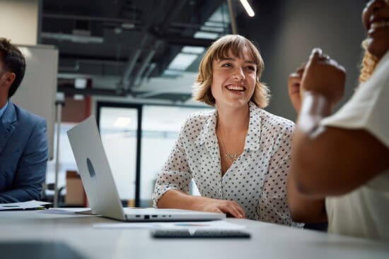 Woman smiling at a meeting table with a laptop, interacting with colleagues in a modern office.