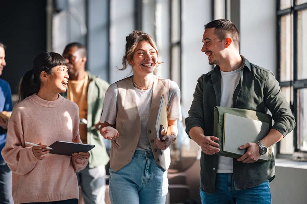 Three young adults walk indoors, smiling and talking, carrying notebooks and a tablet.