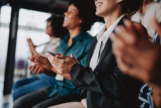 People sitting in a row clapping, shown in a close-up side view at an indoor event or presentation.
