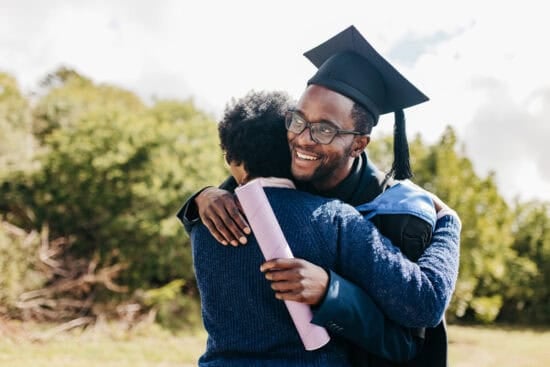 A smiling graduate in cap and gown hugs someone outdoors while holding a diploma.
