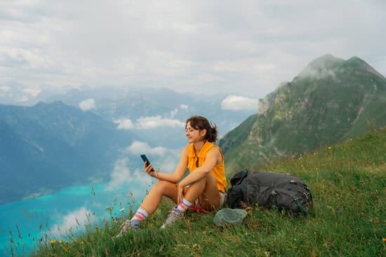 Woman sitting on grassy mountain slope, looking at phone, with backpack and lake view in the background.