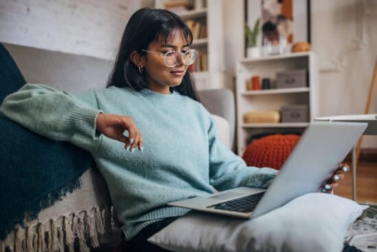 Woman in glasses and a blue sweater sitting on the floor, using a laptop in a cozy living room.
