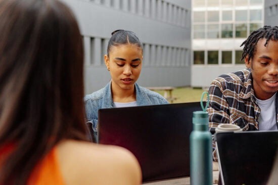 Three students sitting outdoors, working on laptops at a table near a modern building.