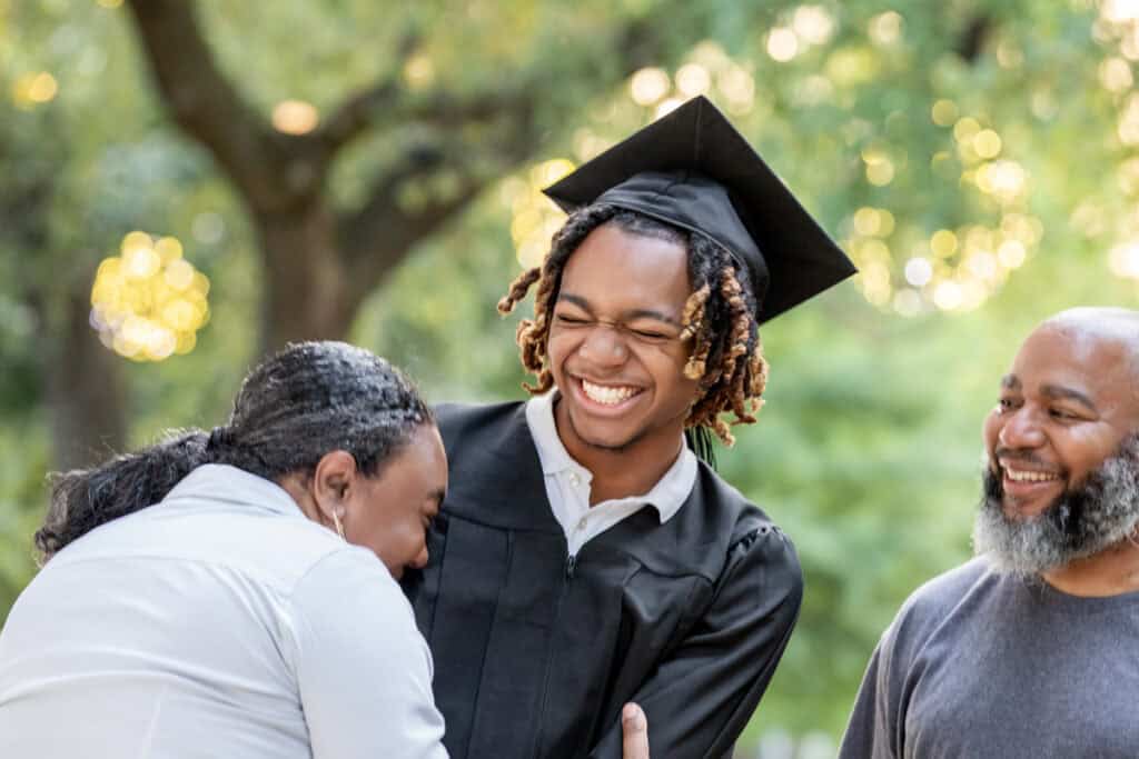 Young graduate in cap and gown smiling joyfully with two happy adults, likely family, outdoors.