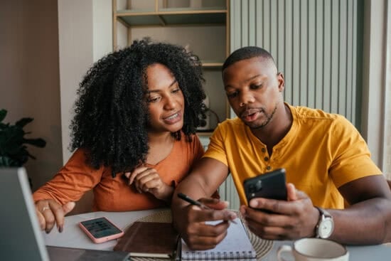 A couple sits at a table, looking at a phone and taking notes together with a laptop nearby.