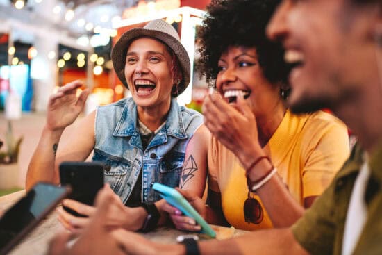 Three friends laughing together at an outdoor table, holding smartphones and enjoying a lively atmosphere.