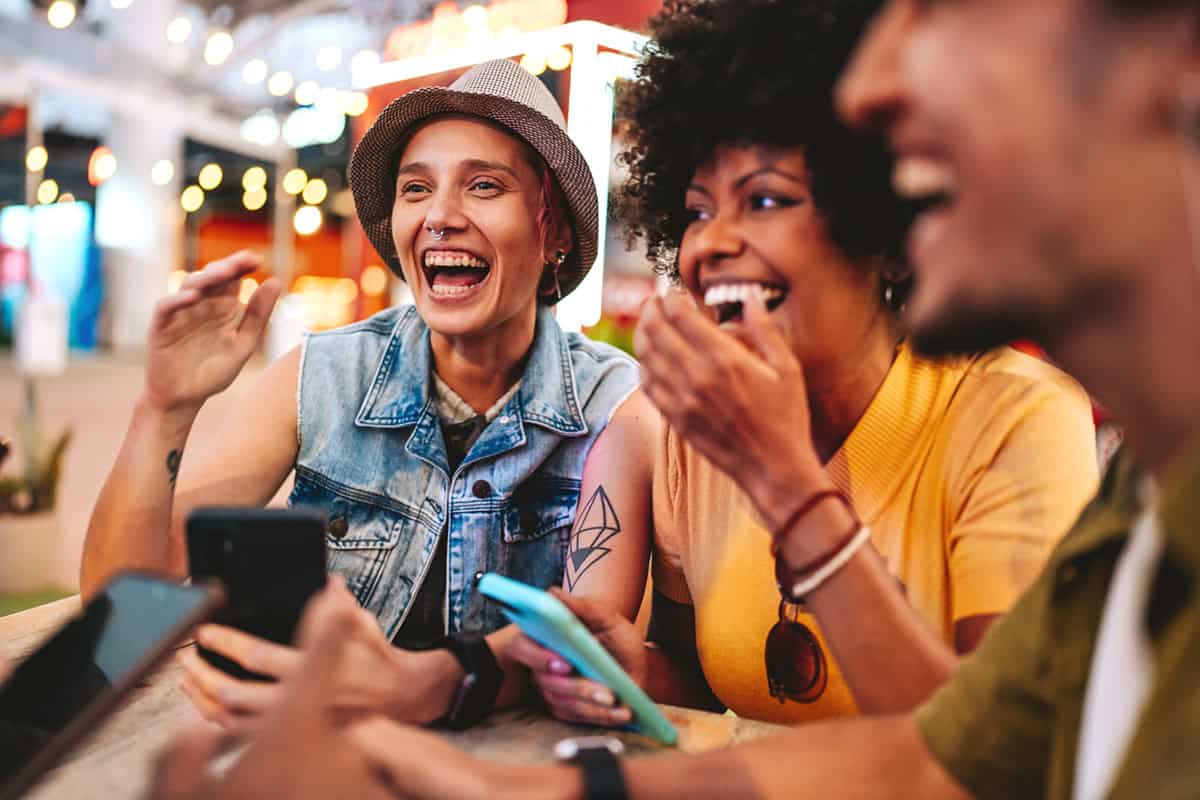 Three friends laughing together at an outdoor table, holding smartphones and enjoying a lively atmosphere.