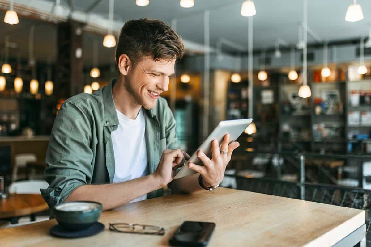 Smiling man using a tablet at a café table with a coffee, glasses, and phone nearby.