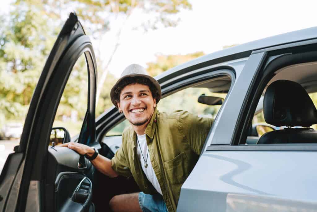 Young man in a hat smiling as he gets out of a car with the door open, outdoors on a sunny day.