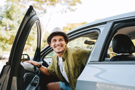 Young man in a hat smiling as he gets out of a car with the door open, outdoors on a sunny day.