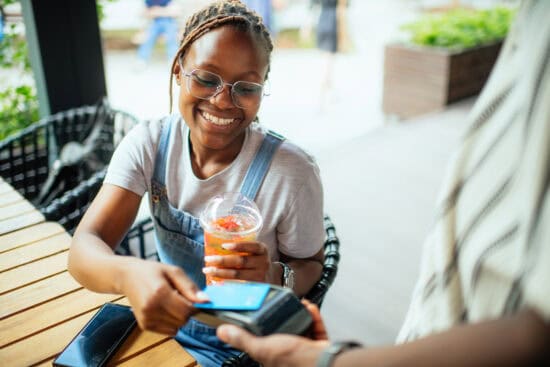 Smiling woman pays with her phone at a cafe while holding a drink in her other hand.