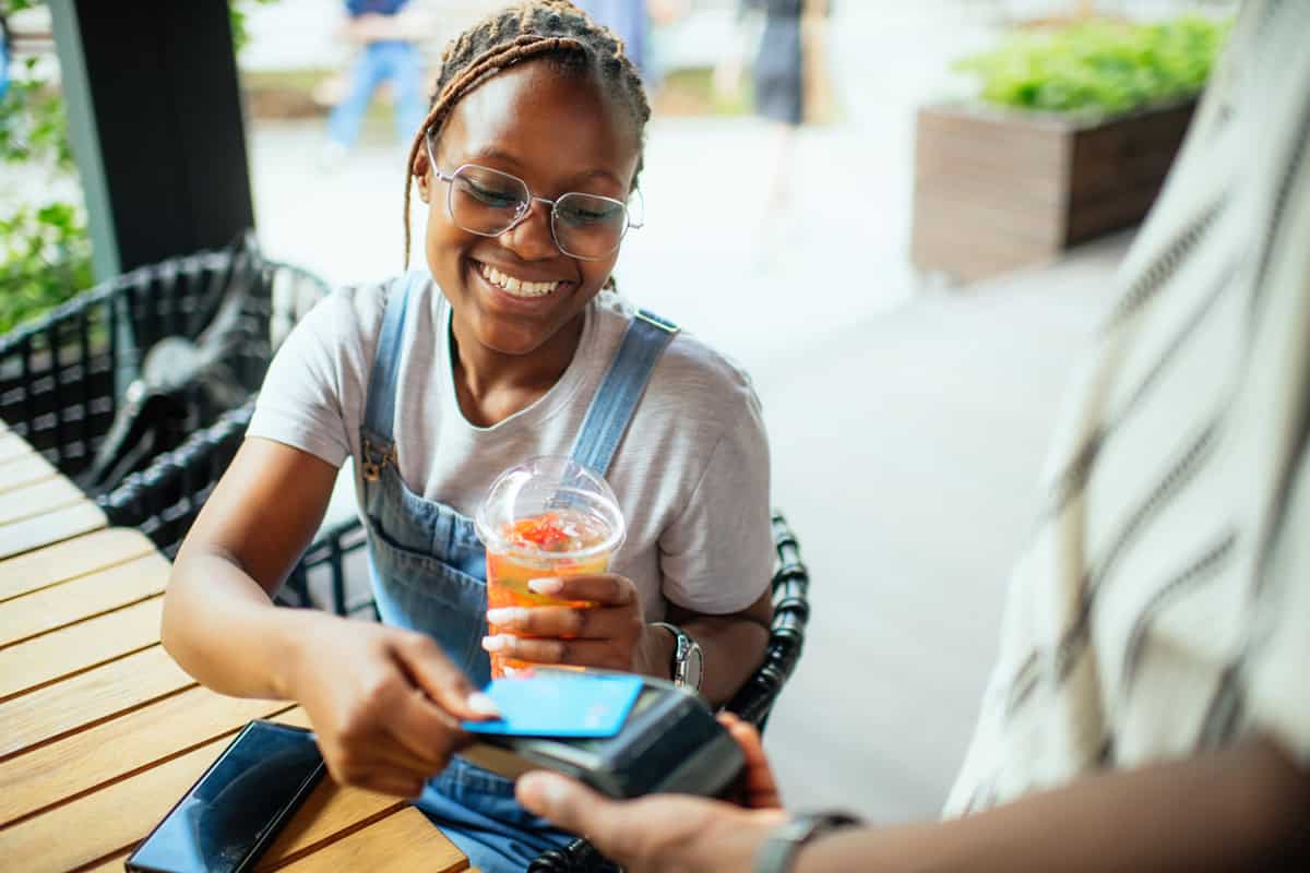 Smiling woman pays with her phone at a cafe while holding a drink in her other hand.