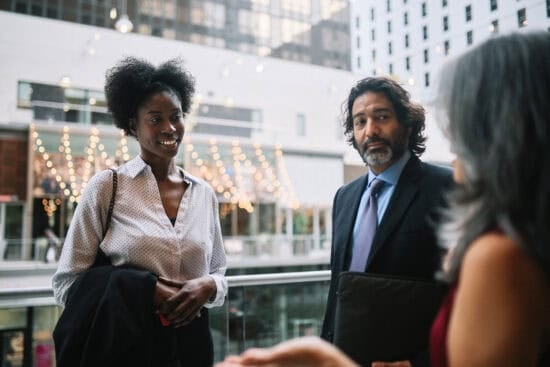Three people in business attire have a conversation on a city balcony with lights in the background.