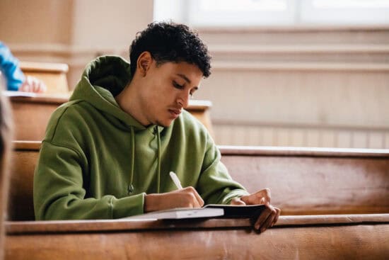 A student in a green hoodie writes in a notebook while sitting in a lecture hall.