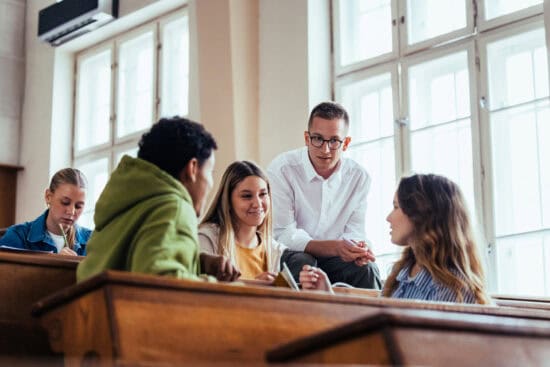A group of students sit and talk together in a sunlit classroom with large windows.