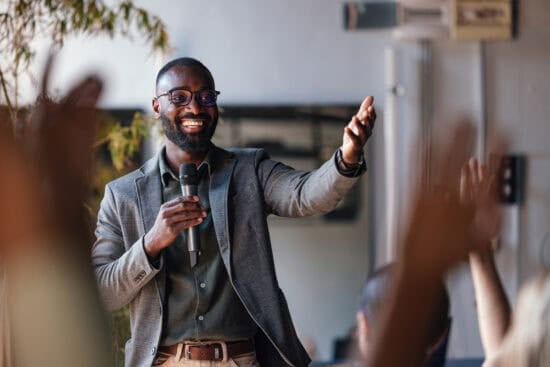 Smiling man in a suit holding a microphone, gesturing to an audience with raised hands.
