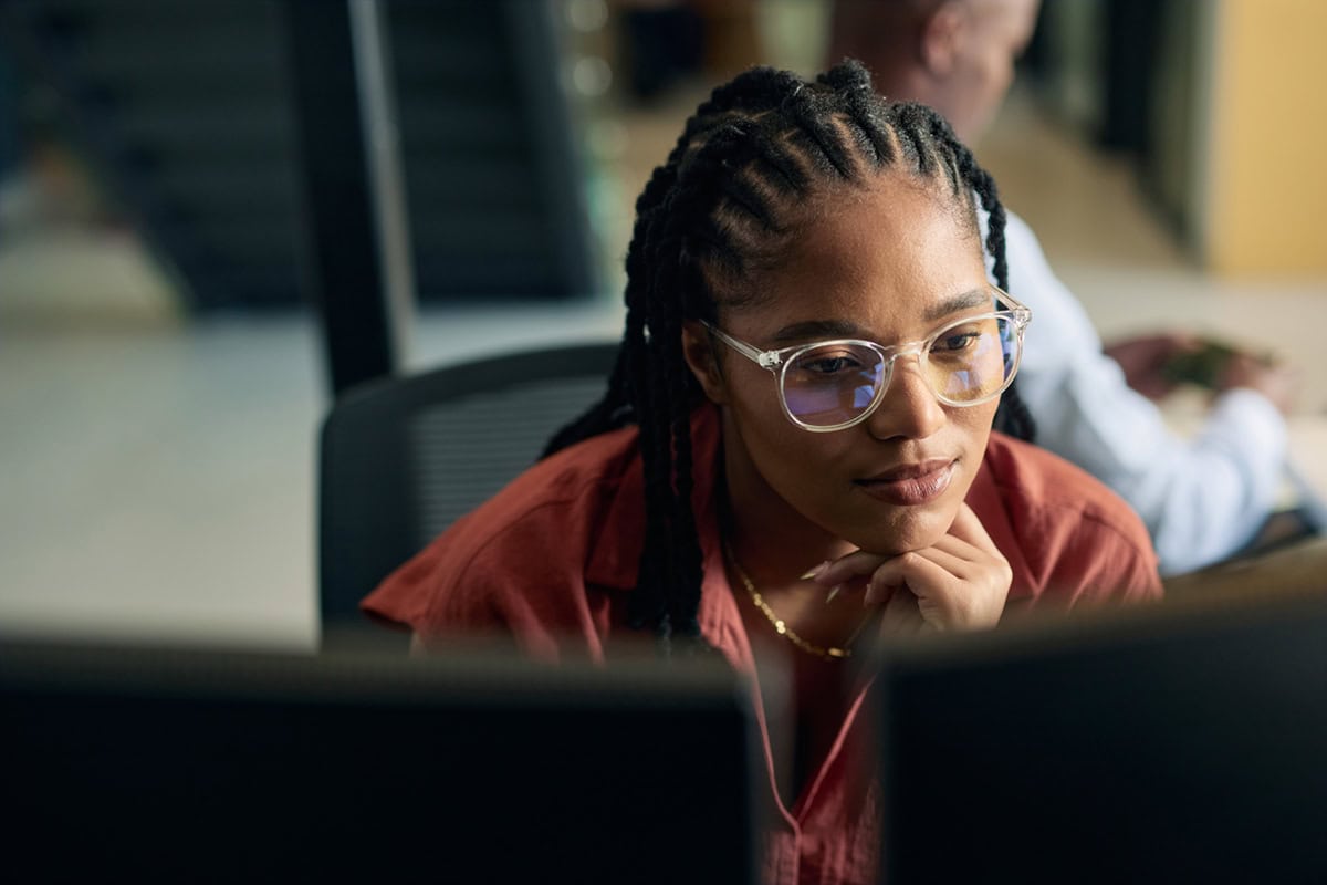 Woman with glasses and braids focuses on a computer screen in an office setting.
